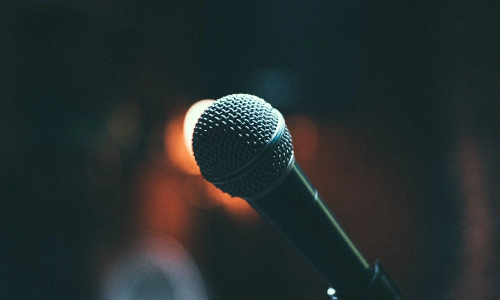 Close-up of a handheld vocal microphone on a stand, illuminated against a dark background with soft, out-of-focus stage lights.