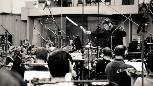 Black and white photo of a conductor wearing headphones leading a full orchestra during a professional recording session in a large studio space with overhead microphones