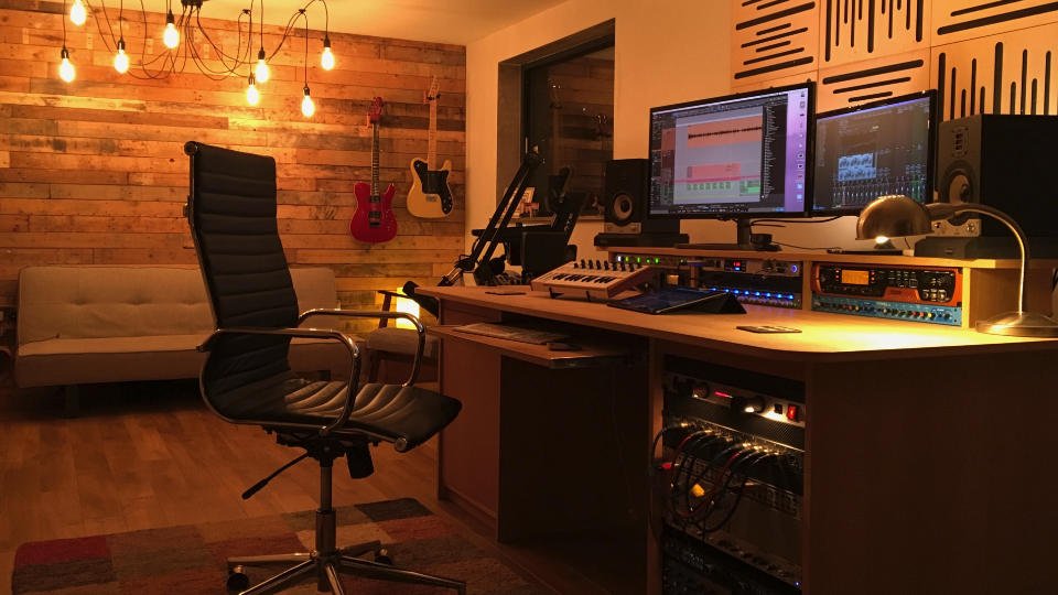 Warmly lit music recording studio with dual monitors, audio mixing equipment, guitars mounted on a wooden wall, and a swivel chair at the production desk.