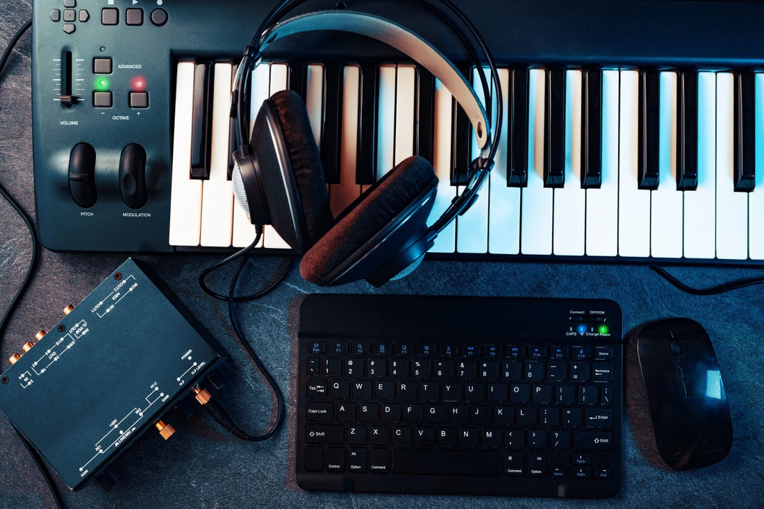 Flat lay of a home studio setup featuring a MIDI keyboard controller, over-ear headphones, a small audio interface, a wireless keyboard, and a computer mouse on a dark textured surface