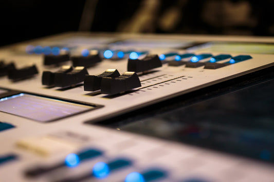 Close-up view of a professional audio mixing console with illuminated blue LEDs, showing multiple faders and control knobs in a studio environment.