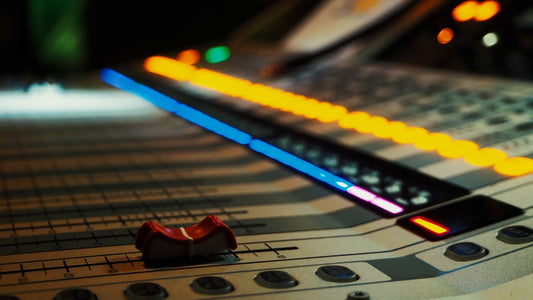 Close-up of a professional audio mixing console with illuminated faders, buttons, and colorful LED lights in a studio environment.