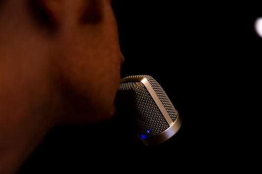 Close-up side view of a person speaking or singing into a studio microphone, with the microphone in sharp focus against a dark, low-light background.