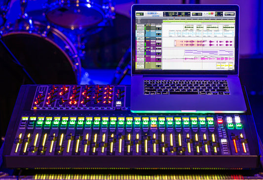 Laptop displaying audio mixing software placed on top of a digital mixing console with illuminated faders and knobs, set in a music studio with a drum kit in the background under blue and purple lighting.