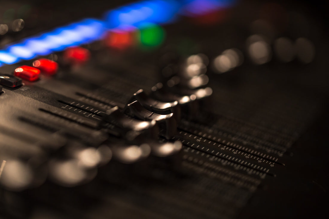 Shallow-focus close-up of an audio mixing console with faders and knobs in the foreground, illuminated by blurred blue, red, and green LED lights in a dark studio setting.