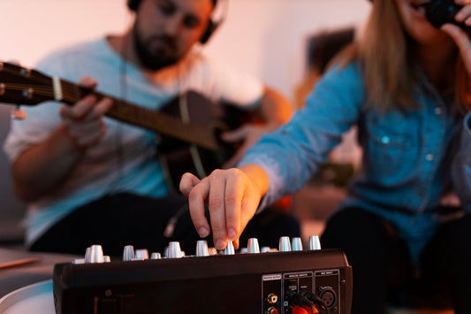 Musicians adjusting audio mixer levels during a home recording session, with one playing guitar and another singing into a microphone, symbolizing collaborative music production.