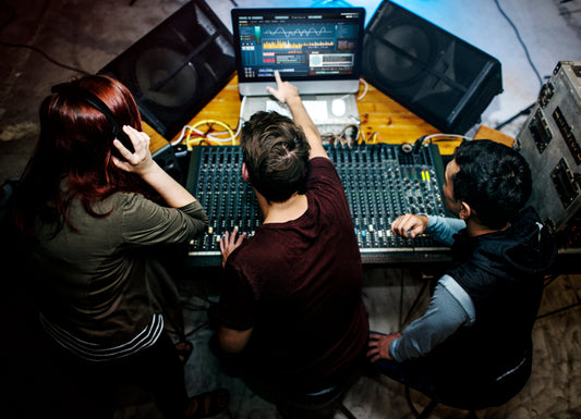 Three people working together at a professional audio mixing console, with one person pointing at a laptop screen displaying sound waveforms and audio data, surrounded by speakers and studio equipment.