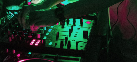 Close-up of a DJ's hands operating a DJ controller with faders and knobs illuminated by green and pink neon lights in a dark performance setting