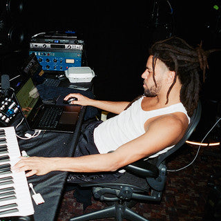 A man with dreadlocks wearing a white tank top sits in a studio chair, working on music production with a laptop, MIDI keyboard, and audio equipment in front of him.