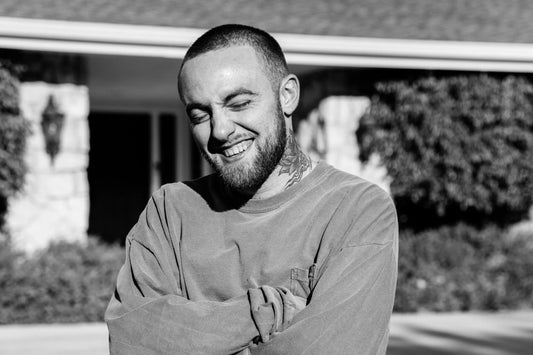 Mac smiling with eyes closed, wearing a sweatshirt and standing outdoors in front of a house, captured in black and white