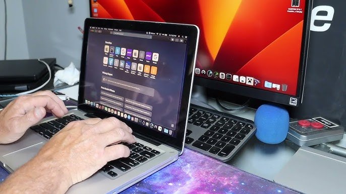 Person typing on a MacBook laptop connected to an external monitor on a desk workspace.