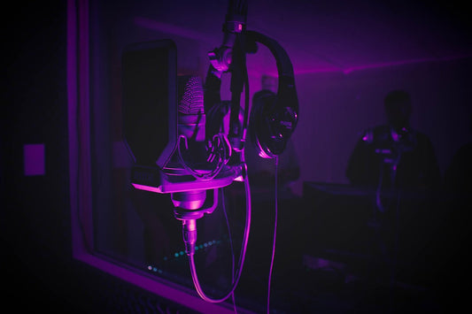 Close-up of a studio condenser microphone with a pop filter and headphones hanging on it, illuminated by purple lighting inside a recording booth, with blurred figures in the background.