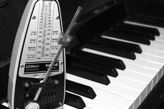 Black-and-white close-up of a mechanical metronome positioned in front of piano keys, with the tempo scale visible and the pendulum slightly blurred in motion.