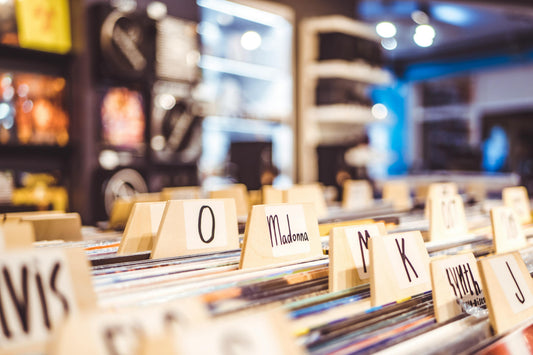 Record store vinyl bins with wooden alphabet dividers, including a visible “Madonna” label, in a softly blurred music shop interior.