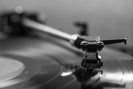 Close-up black and white image of a turntable with a tonearm and Shure cartridge resting on a spinning vinyl record, highlighting the grooves of the record.