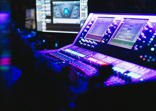 A professional audio engineer operating a large digital mixing console with glowing faders and knobs, illuminated by purple and blue stage lighting with DAW software visible on monitors in the background