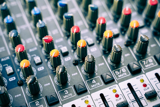 Close-up of an audio mixing console with multiple rotary knobs in different colors, labeled channels, pan controls, mute buttons, and LED clip indicators.
